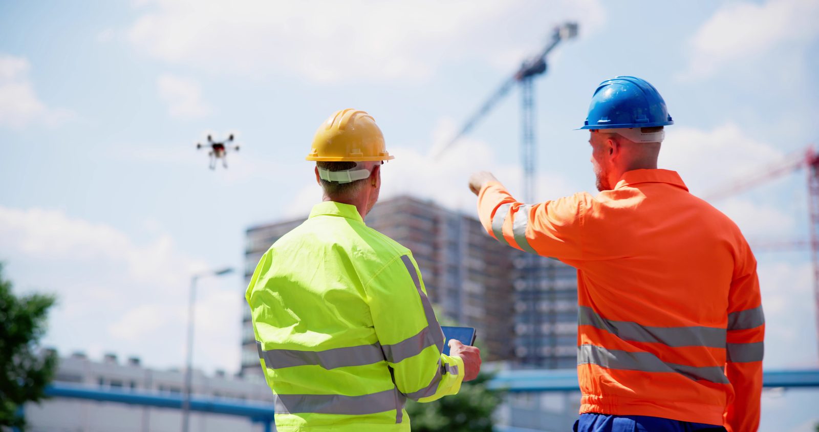 Dva pracovníci v reflexních vestách sledují dron nad staveništěm